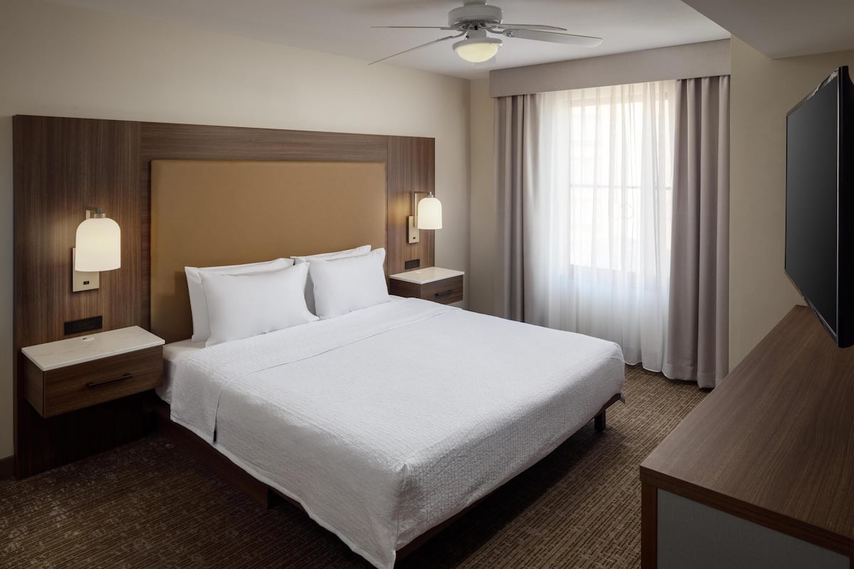 Una habitación de hotel con una cama grande, dos mesitas de noche con lámparas, un ventilador de techo, un ventanal con cortinas y un televisor de pantalla plana.