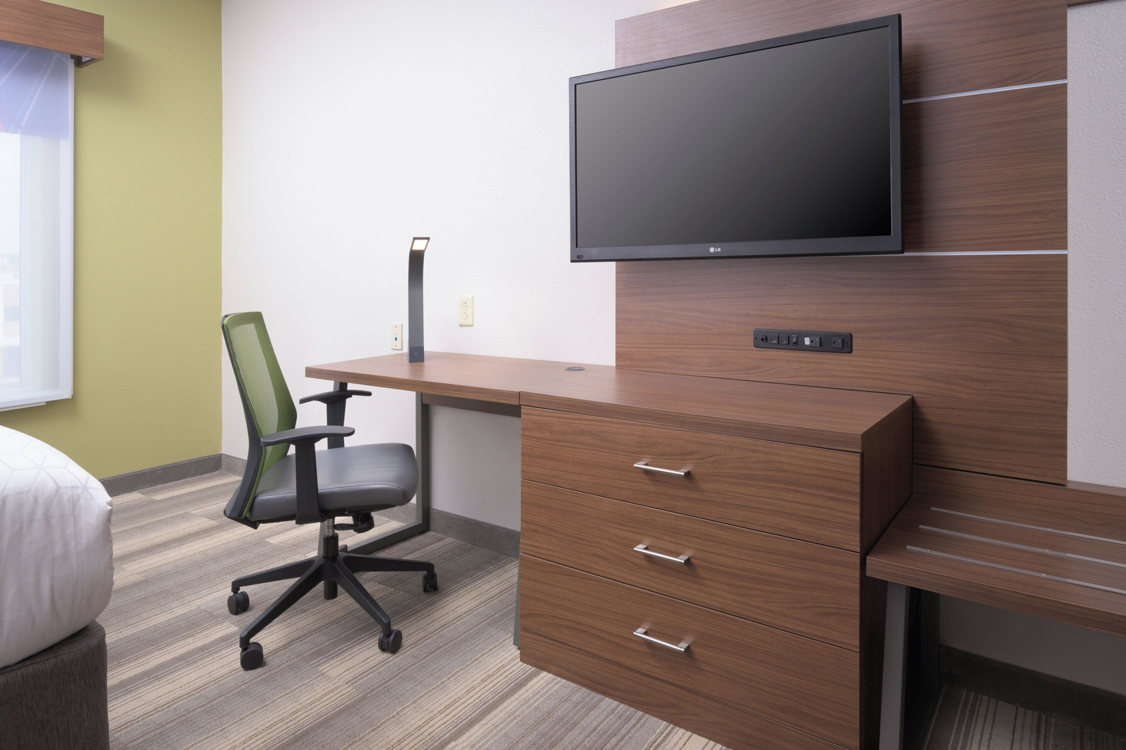 A hotel room with a wooden desk, a modern chair, a flat-screen TV mounted on the wall, and a bed with a white pillow.
