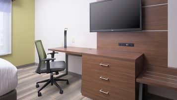 A hotel room with a wooden desk, a modern chair, a flat-screen TV mounted on the wall, and a bed with a white pillow.