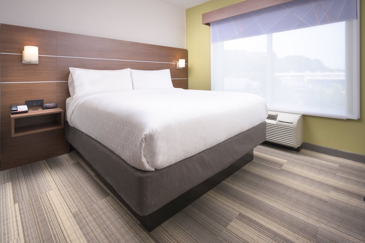 Une chambre d’hôtel moderne dotée d’un grand lit, d’une table de chevet avec un téléphone et un chargeur, d’une fenêtre avec des stores et d’un radiateur fixé au mur.