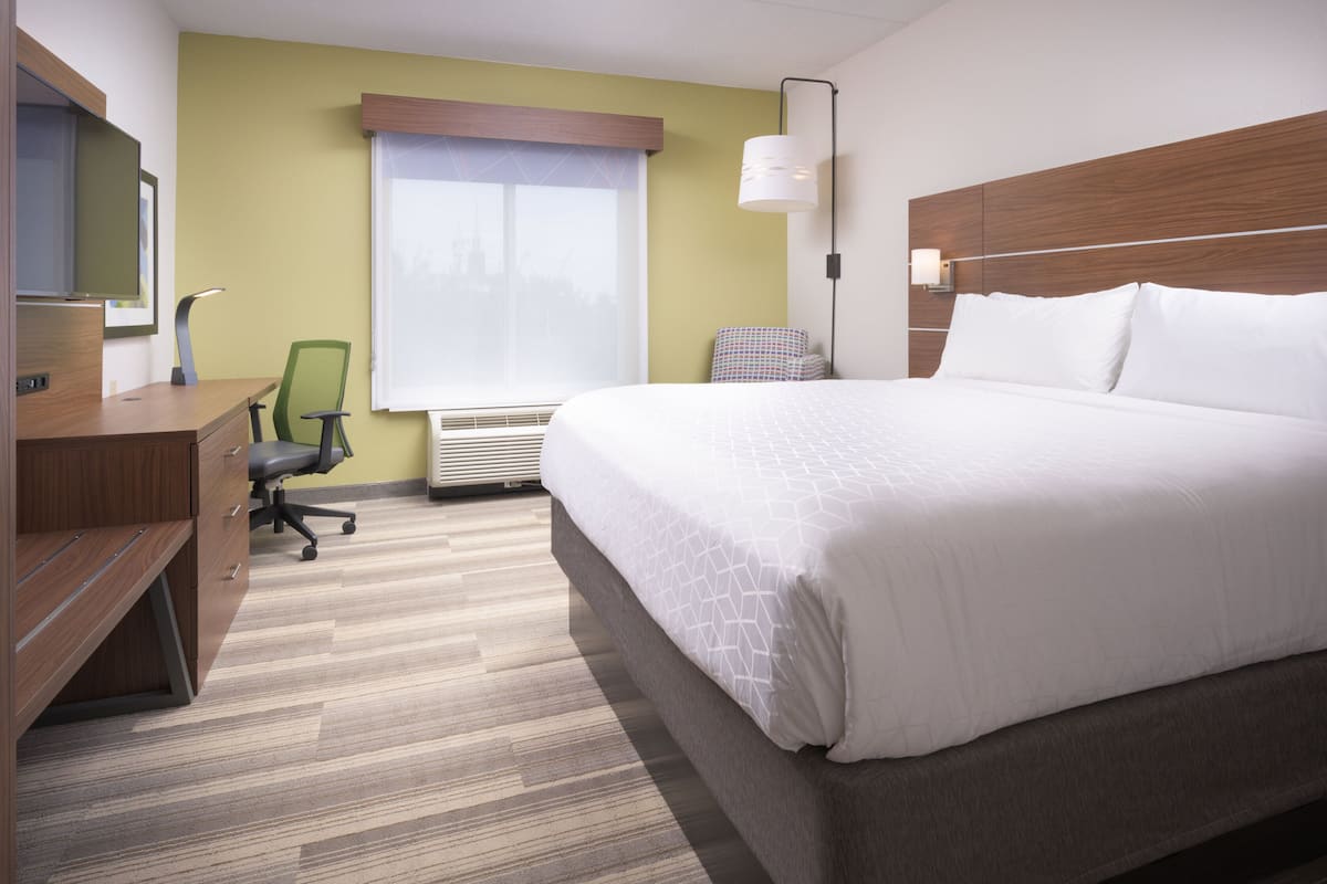 Une chambre d’hôtel avec un grand lit, un bureau avec une chaise, une fenêtre avec des stores et une lampe fixée au mur.