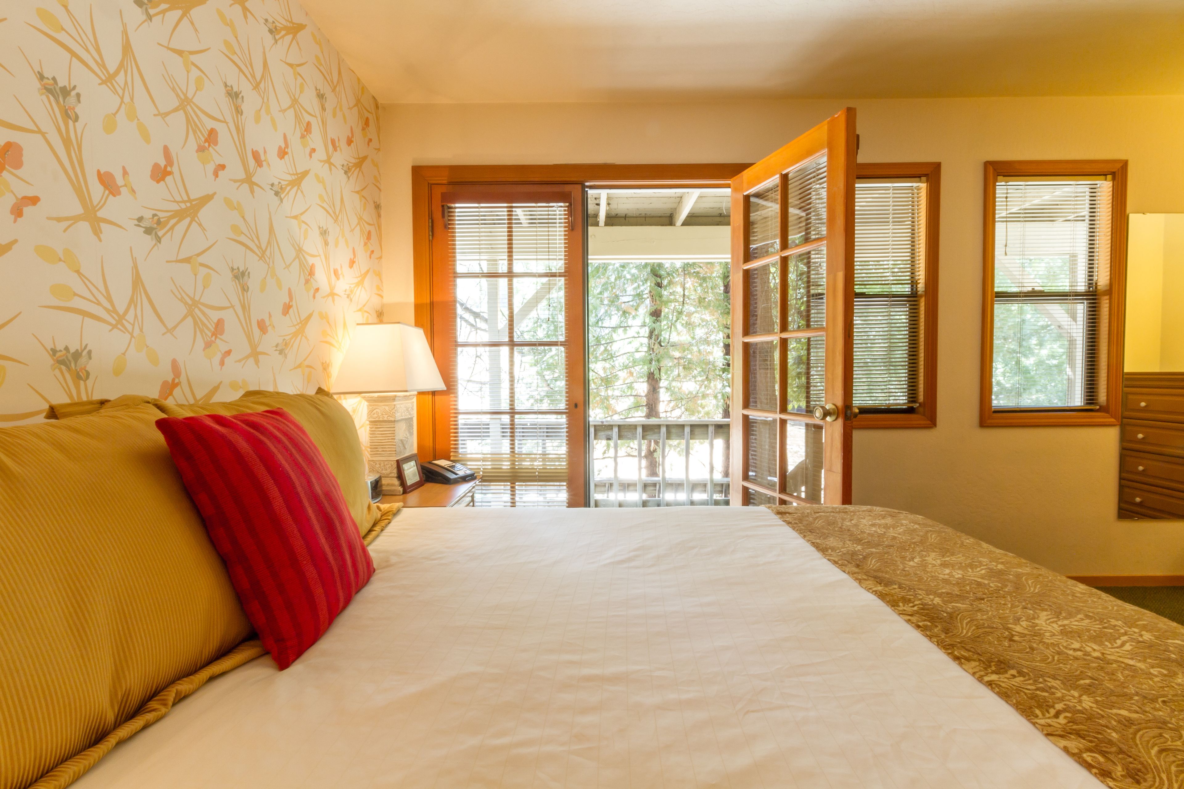 A hotel room with a bed, a nightstand, a lamp, and a view through a door to an outdoor area.