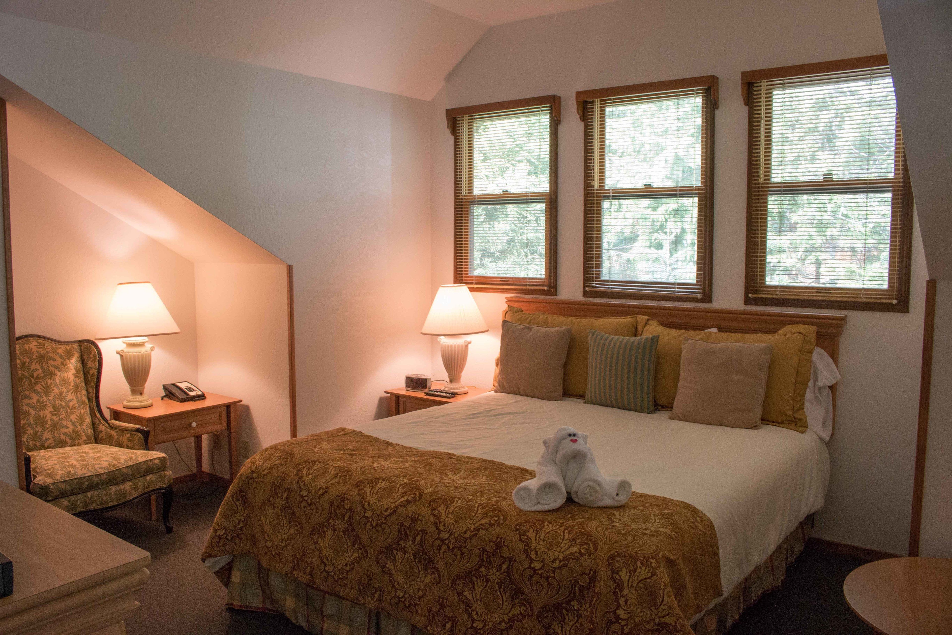 A hotel room with a bed, two bedside lamps, a chair, and a window with blinds.