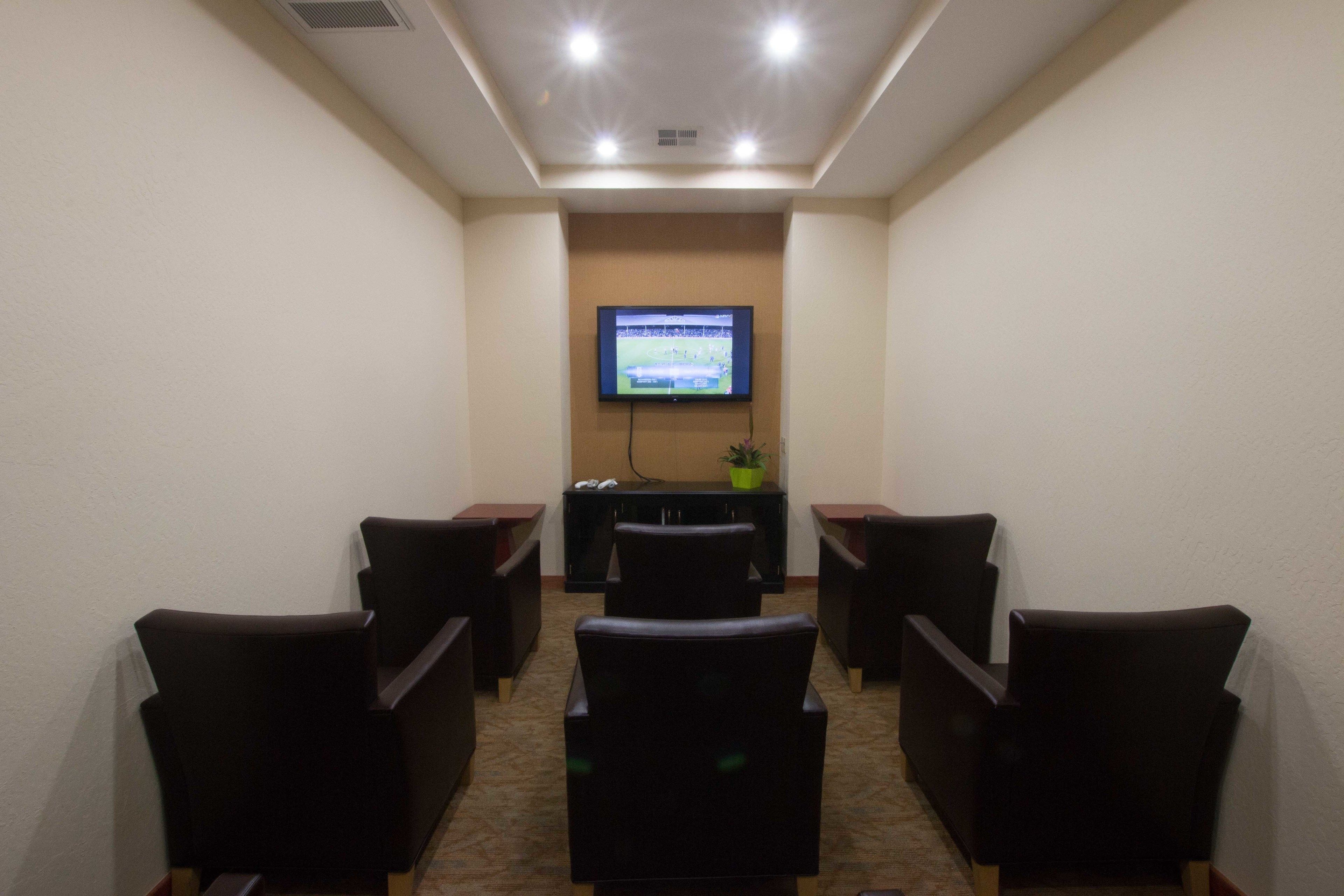 A conference room with a flat-screen TV mounted on the wall, a small table with a potted plant, and six black chairs arranged in two rows.