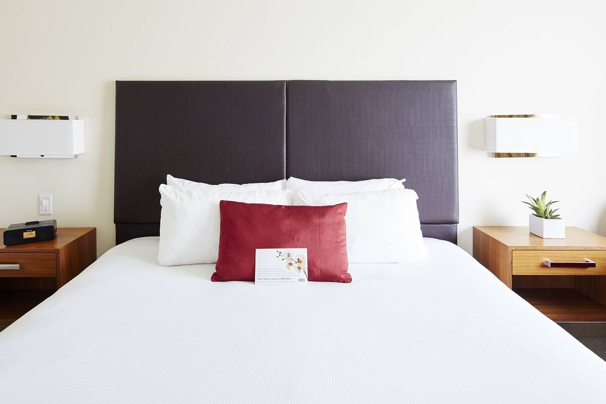 A hotel room with a large bed, two bedside tables, a nightstand, a red chair, a small plant, and a wall-mounted lamp.