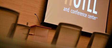 Un écran de projection sur lequel est affiché le texte « HÔTEL et centre de conférences », dans une salle de conférence équipée de chaises.