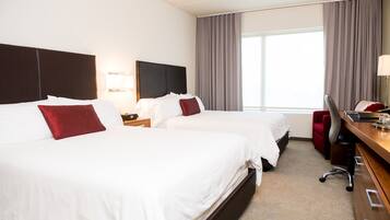 A hotel room with two beds, a desk with a chair, a lamp, and a large window with curtains.