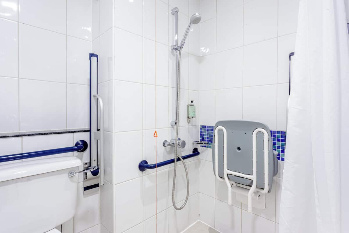 A bathroom with white tiles, a shower with a handheld showerhead, a toilet with a grab bar, and a soap dispenser.