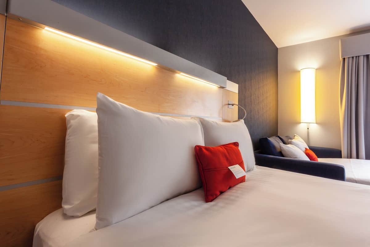 A hotel room with a wooden headboard, a large bed with white and red pillows, a bedside table, and a lamp.