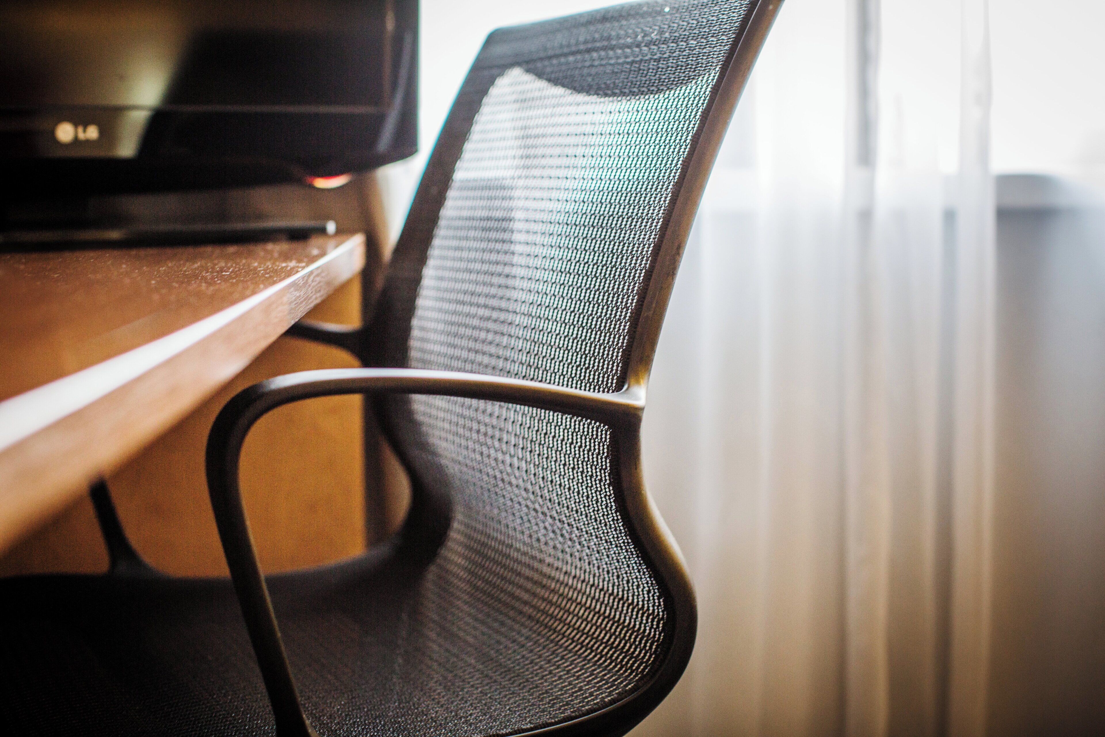 A close-up of a black mesh office chair with a wooden desk and a black monitor.