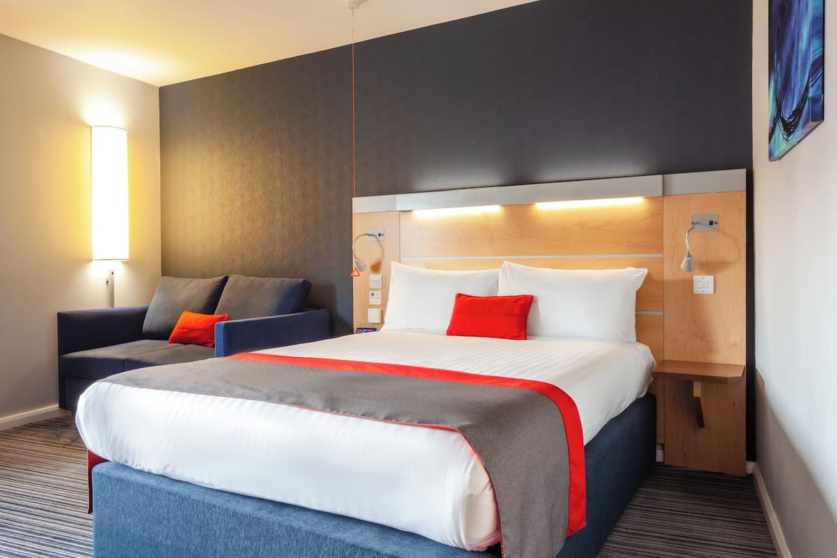 A hotel room with a large bed, a sofa, a bedside table, and a wall-mounted lamp.