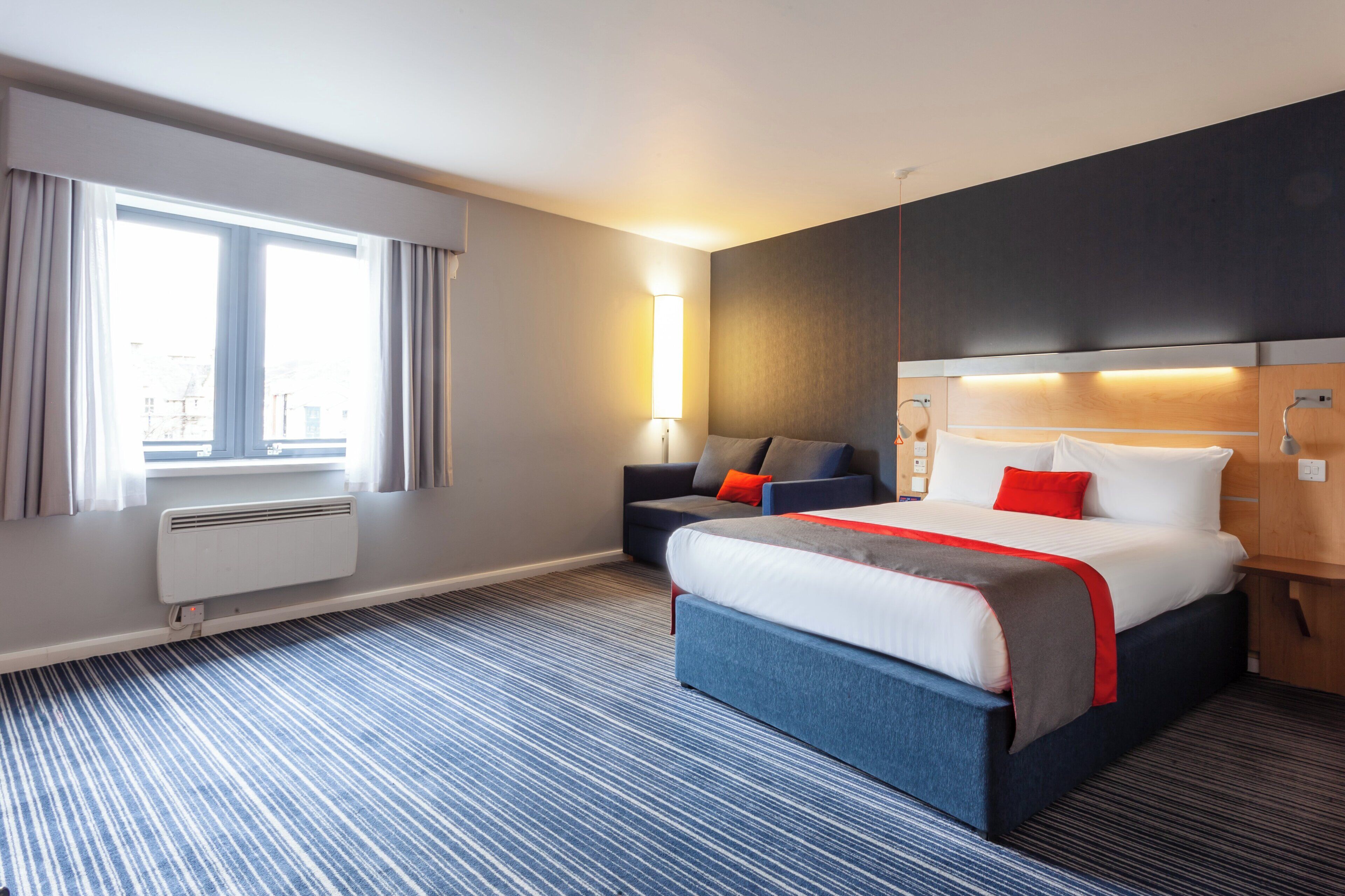 Ett hotellrum med en stor säng, en mindre soffa, ett sängbord, en lampa och ett fönster med gardiner.