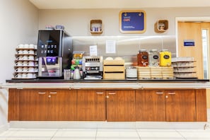 A coffee station with a coffee machine, milk frother, and various coffee pods. There are also jars of water and orange juice, and a sign that says 'Wake Cup!'