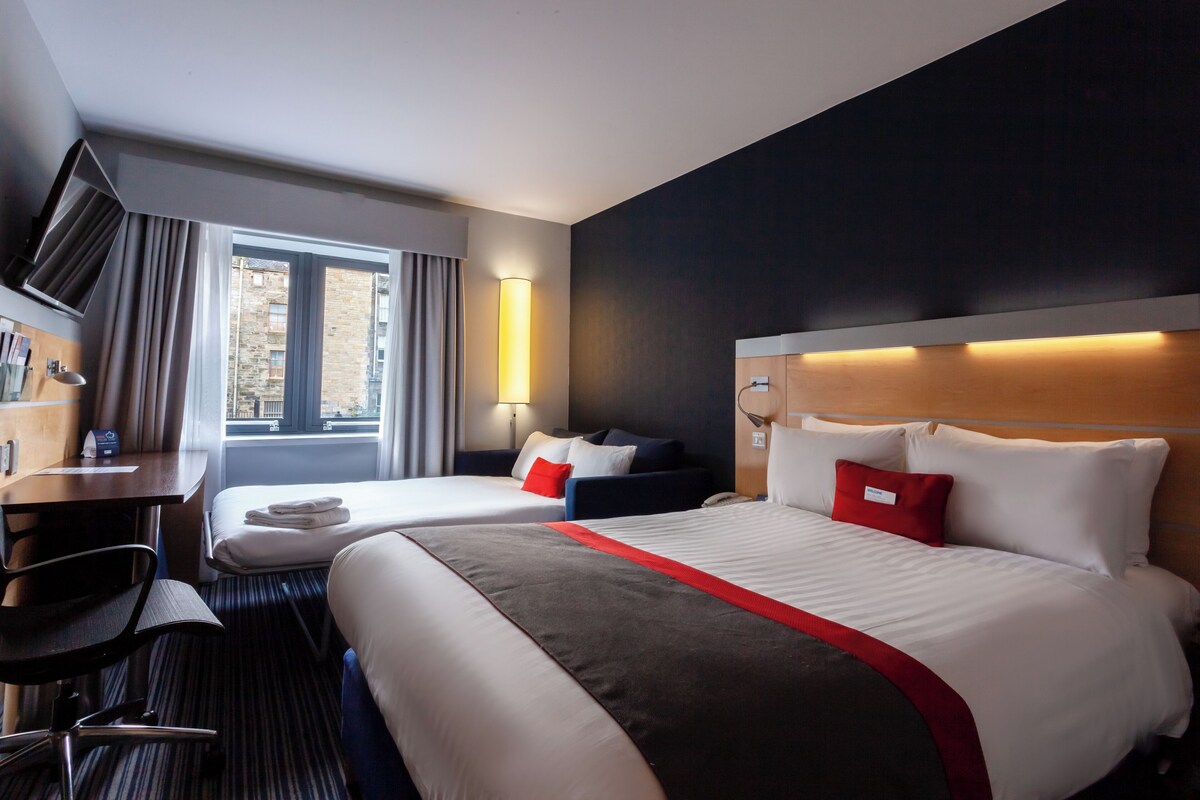 Quarto de hotel com duas camas, uma escrivaninha, uma cadeira, uma televisão e uma janela com cortinas.