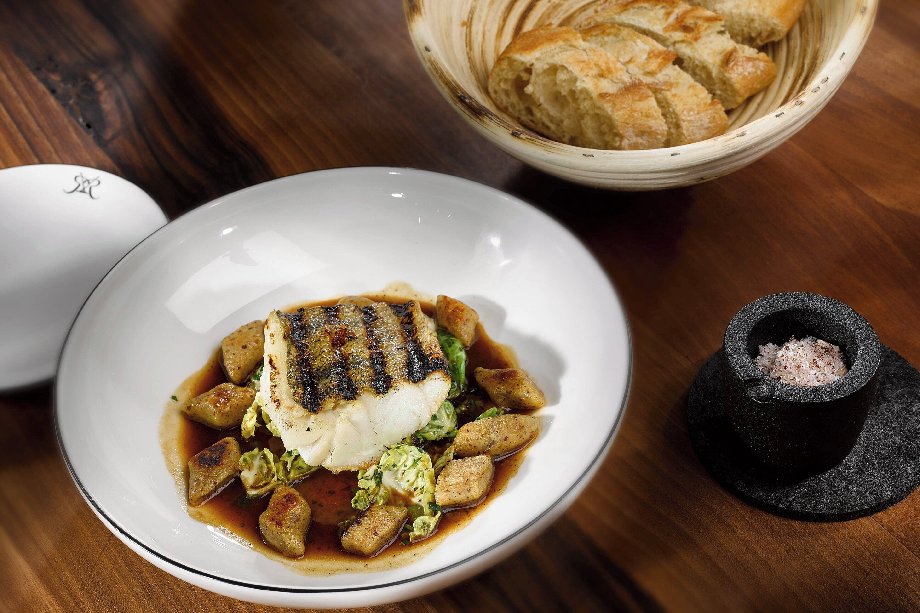 A plate of food with grilled fish, potatoes, and greens, a bowl of bread, and a salt shaker on a wooden table.