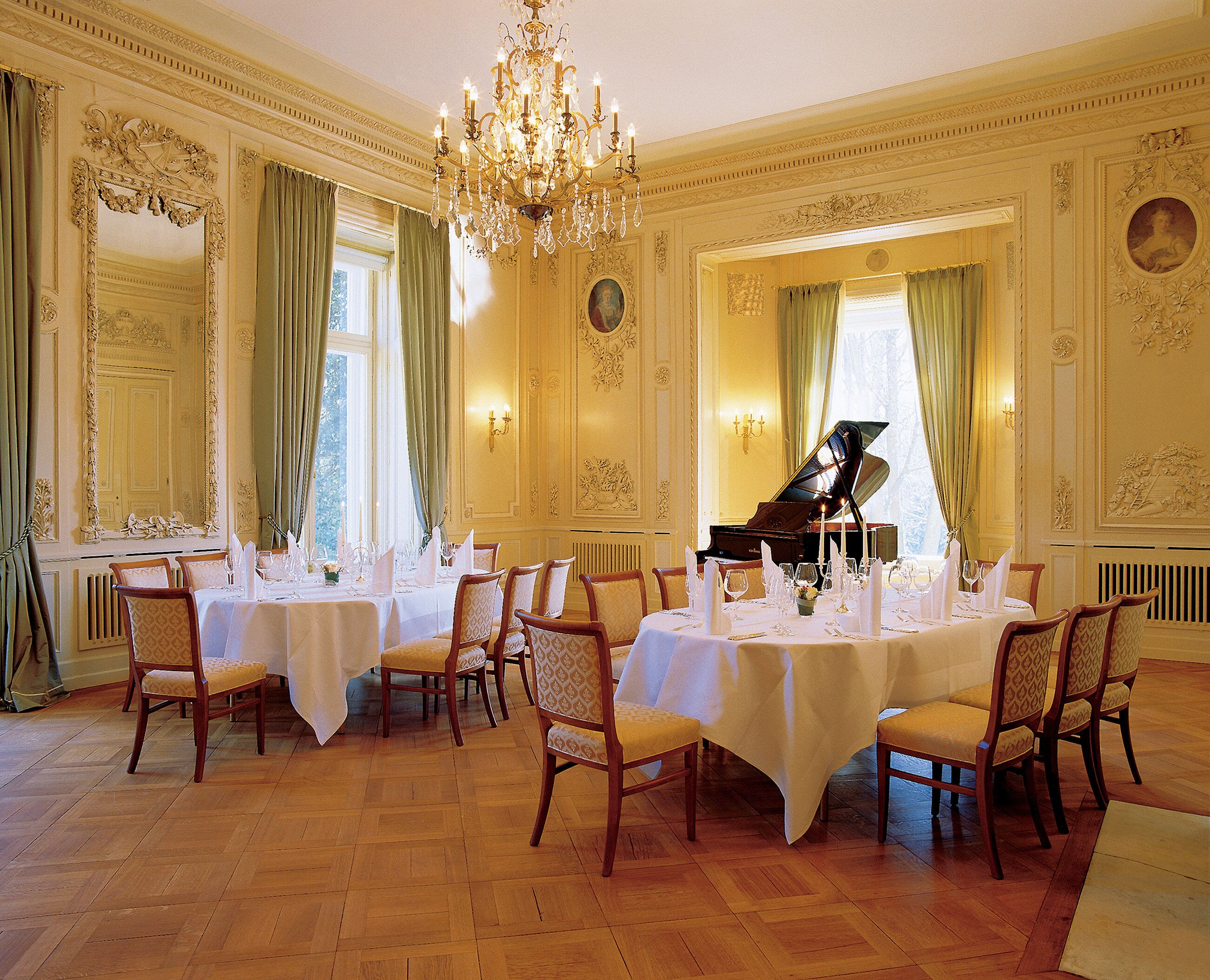 A formal dining room with tables set for a meal, a grand piano, and ornate wall decorations.