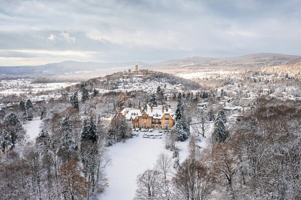 Un grande edificio storico con una torre imponente, circondato da alberi innevati e un paesaggio collinare.