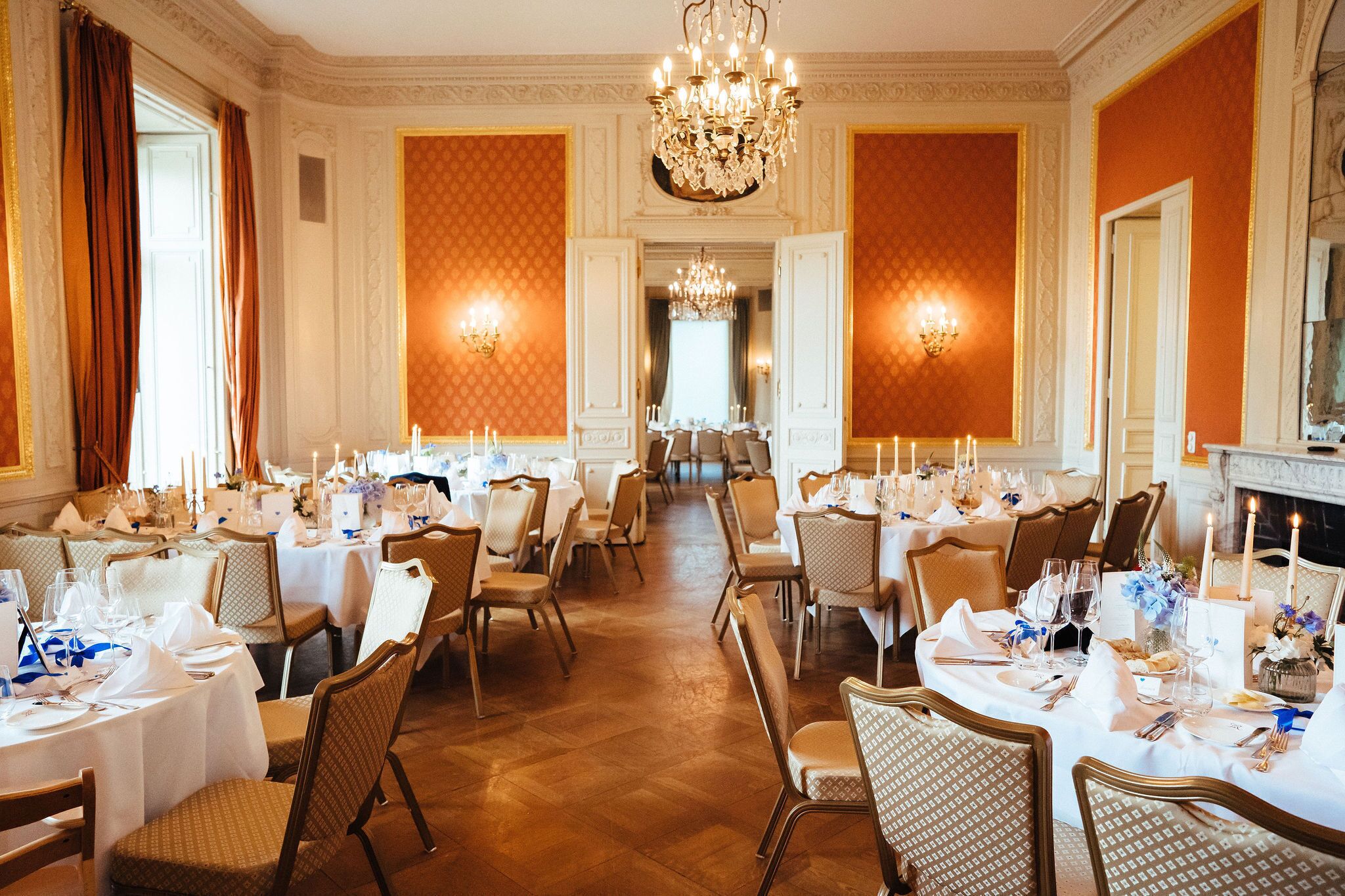 A formal dining room with multiple tables set for a meal, elegant chandeliers, and a fireplace.