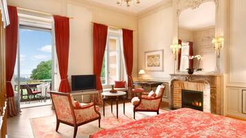 A luxurious hotel room with a large bed, two armchairs, a small table, a fireplace, and a TV.