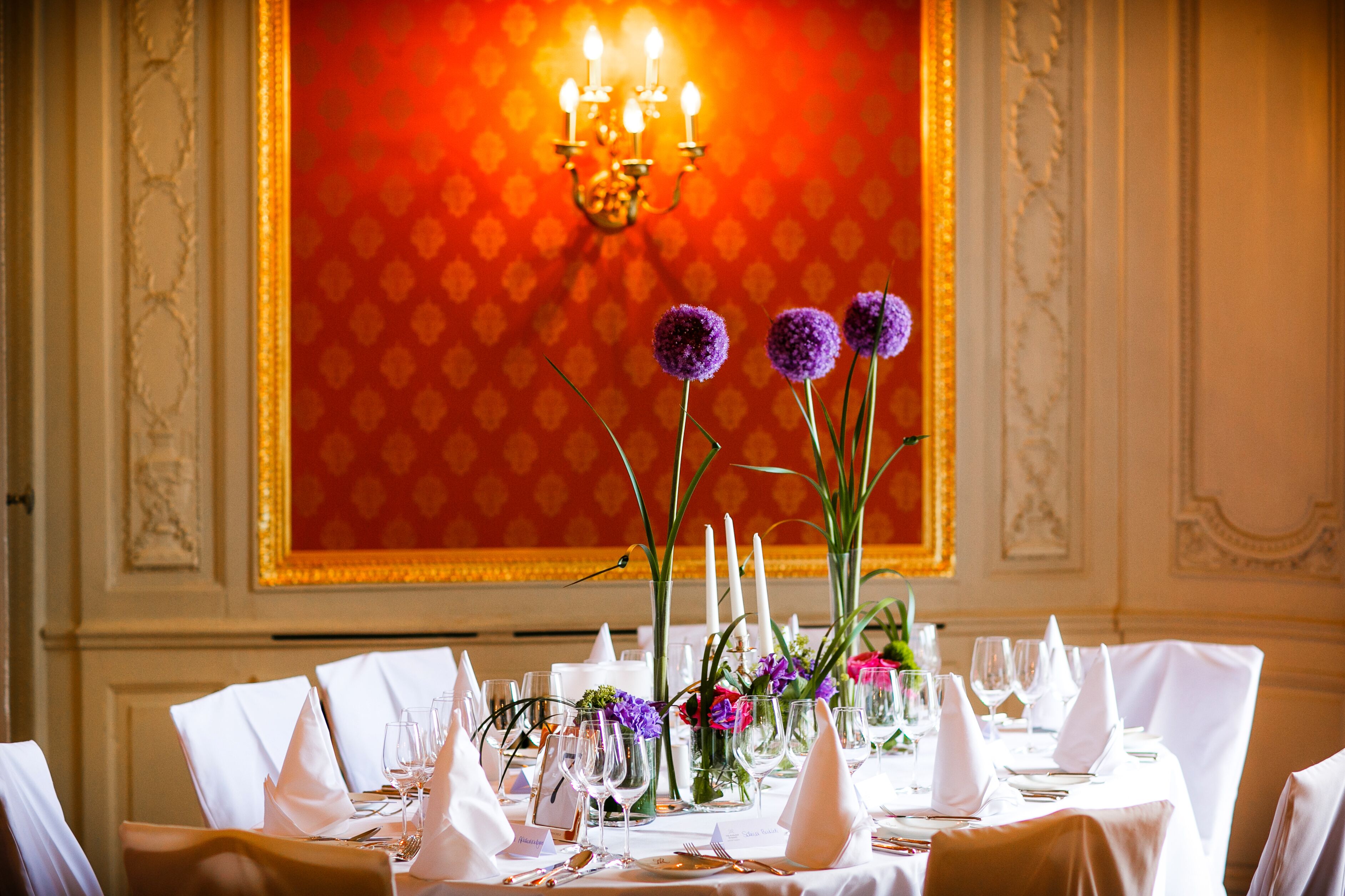 A formal dining table set with white tablecloths, glassware, and purple flowers.