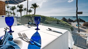 Mesa para dos con cristalería azul, con vista a una playa con palmeras y chozas de techo de paja.