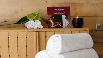 A neatly made bed with rolled white towels and a green blanket, a lit candle, and a book titled "Cinq Mondes Spa Marseille" on a wooden shelf.