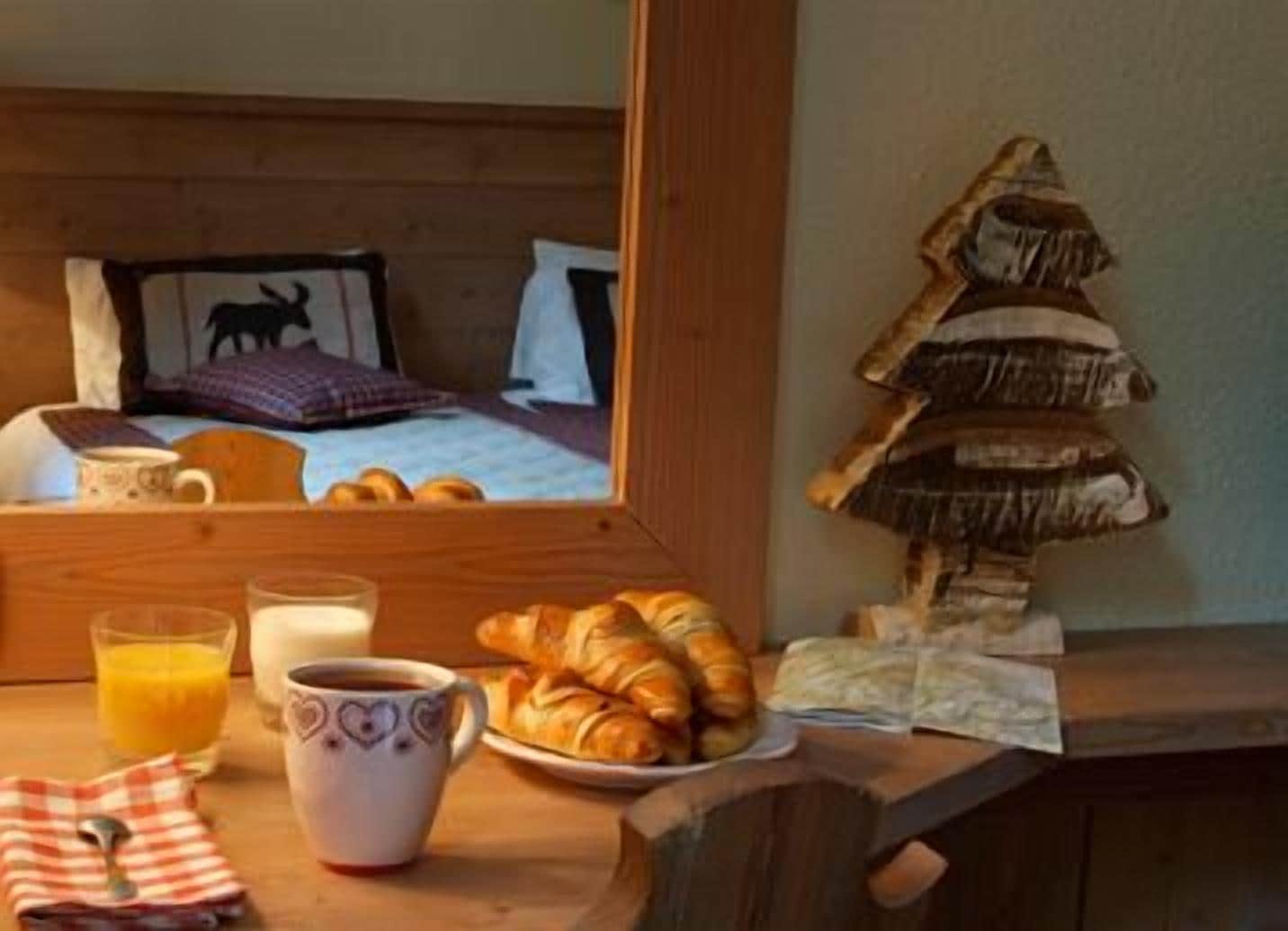 Une pièce chaleureuse avec un lit en bois, un petit arbre décoratif, des croissants, une tasse de café et un verre de jus d’orange sur une table en bois.