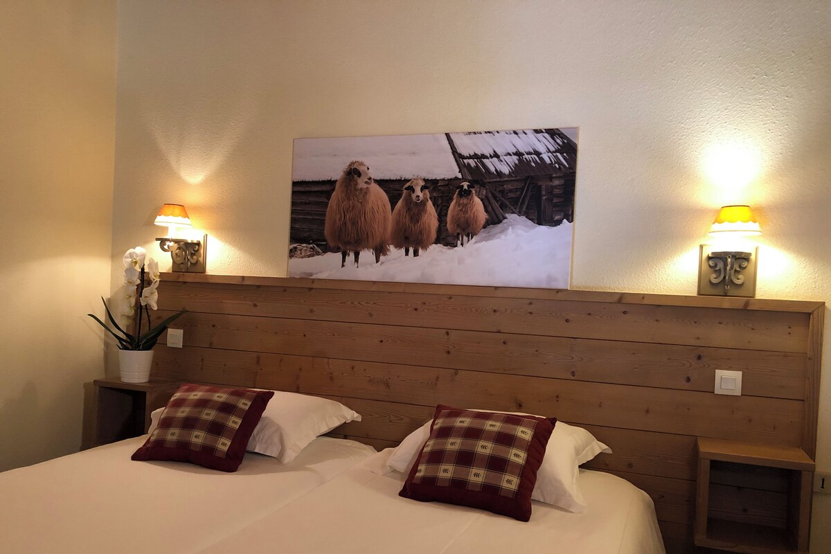 A hotel room with a wooden headboard, two bedside tables with lamps, a bed with white bedding and plaid pillows, a wall-mounted painting of sheep, and a potted orchid.