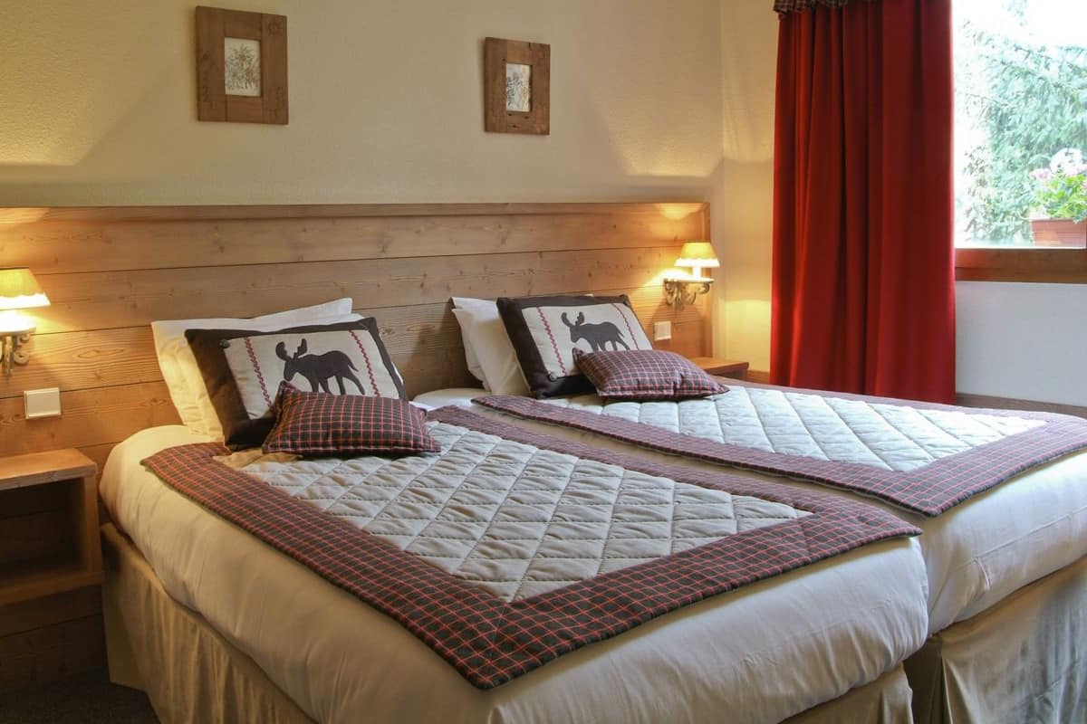 Uma cama de casal com colcha e duas almofadas, uma mesa de cabeceira com abajur, e uma janela com cortinas vermelhas.