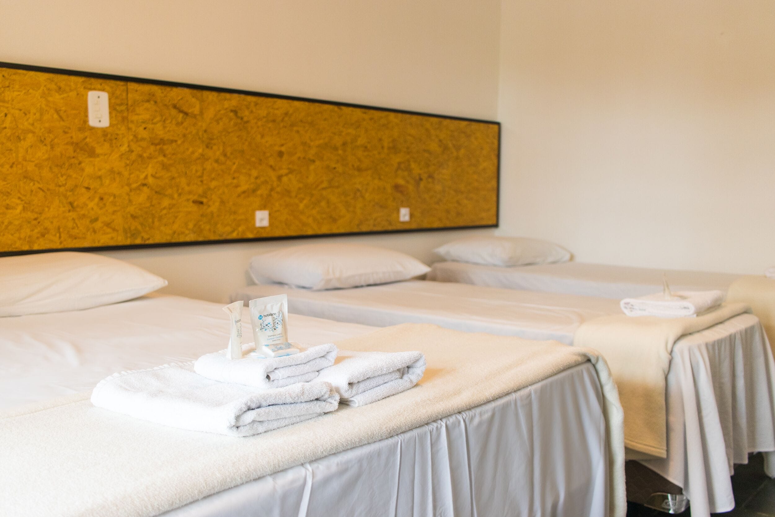 A hotel room with three beds, each with white linens and pillows. There is a small table with a towel and a card on it.