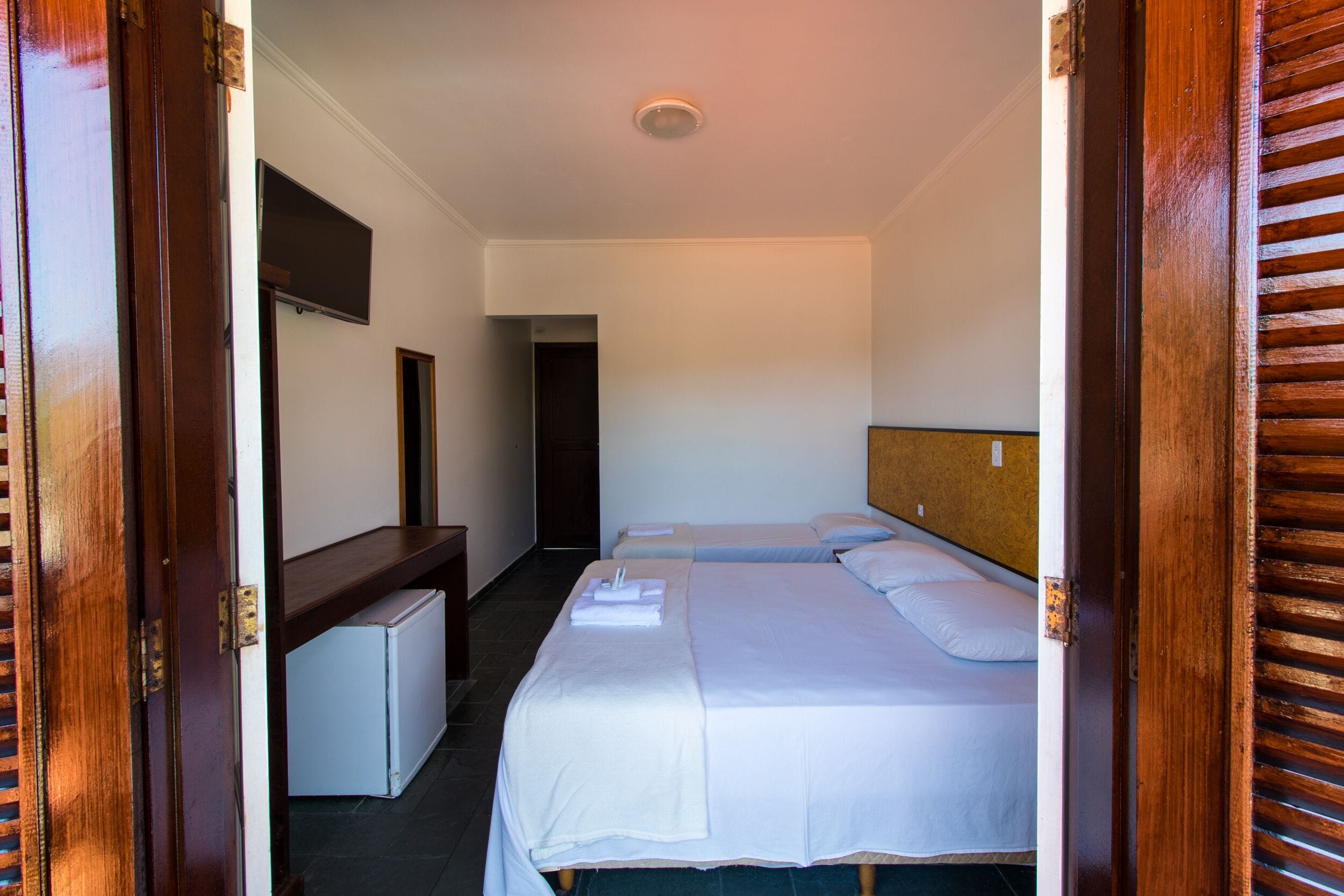 Habitación de hotel con dos camas, un escritorio pequeño, televisor y una puerta con persianas de madera.