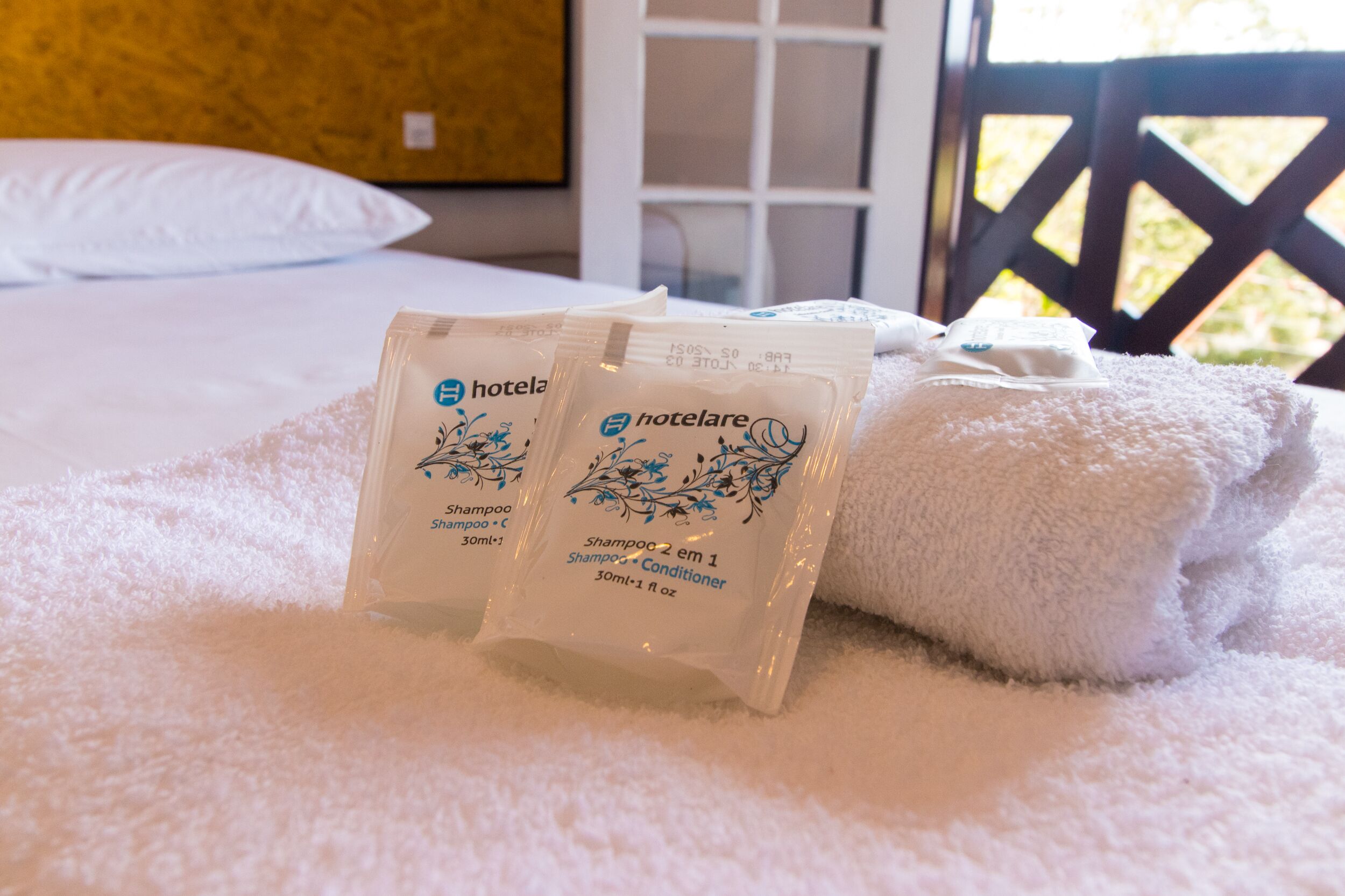 Una cama con sábanas blancas, dos paquetes de artículos de aseo de hotel y una toalla blanca sobre una superficie mullida y blanca.