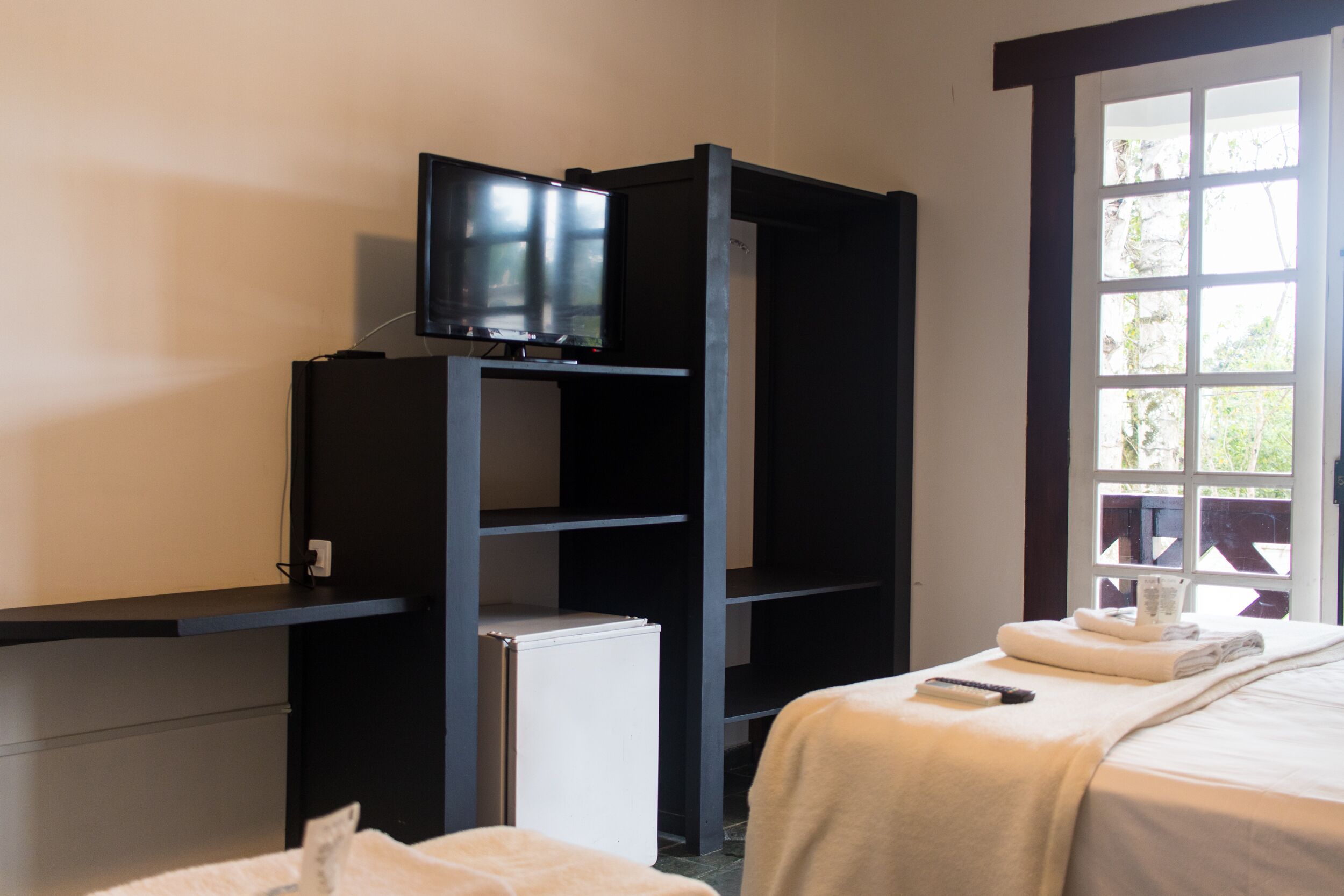 Habitación de hotel con cama, televisor en un estante, refrigerador pequeño y ventana con vista a árboles.