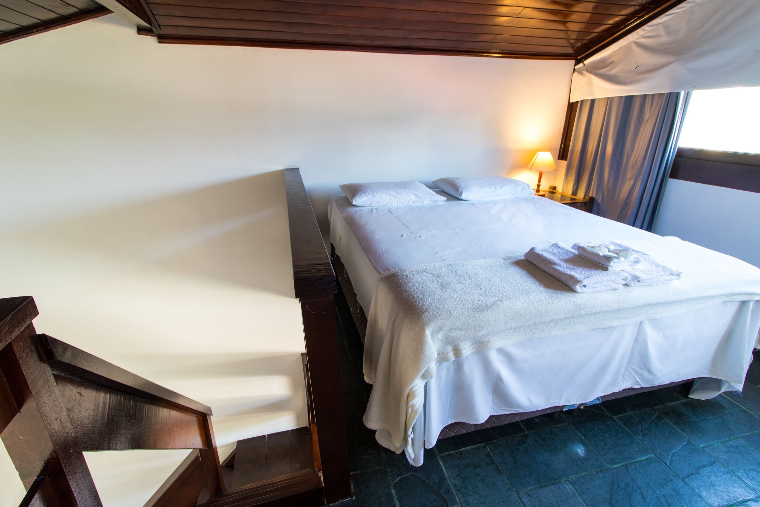 A neatly made bed with white linens and towels, a wooden chair, and a nightstand with a lamp.