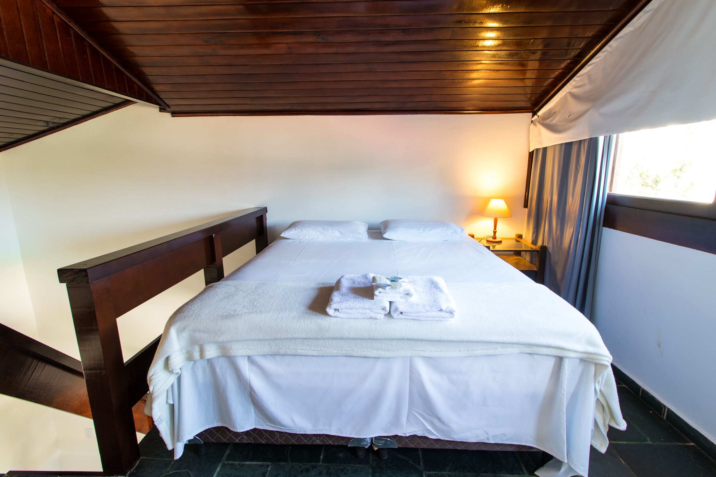 Una cama bien hecha con ropa de cama y toallas blancas, cabecera de madera y una mesita de noche con lámpara.