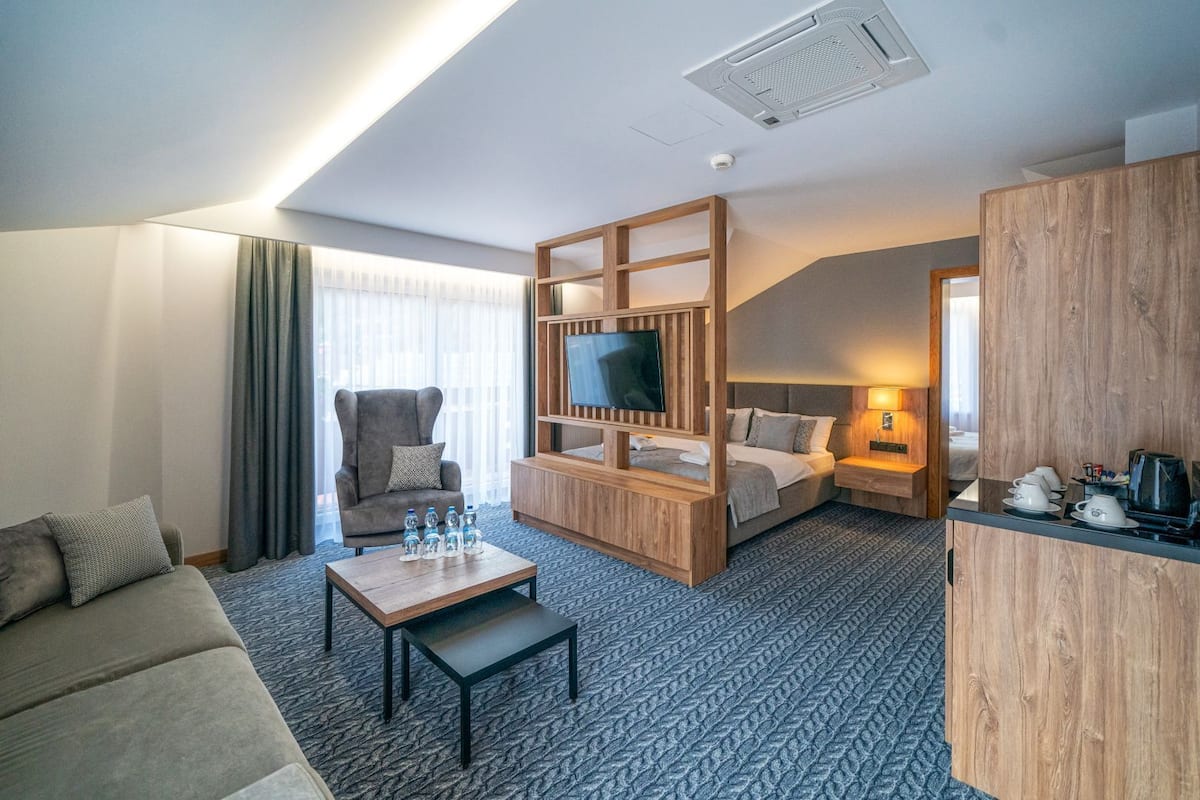 Habitación de hotel con sofá, una mesa de centro, un televisor montado en una pared de madera, una cama con cabecera, una mesita de noche con lámpara y un área de kitchenette.