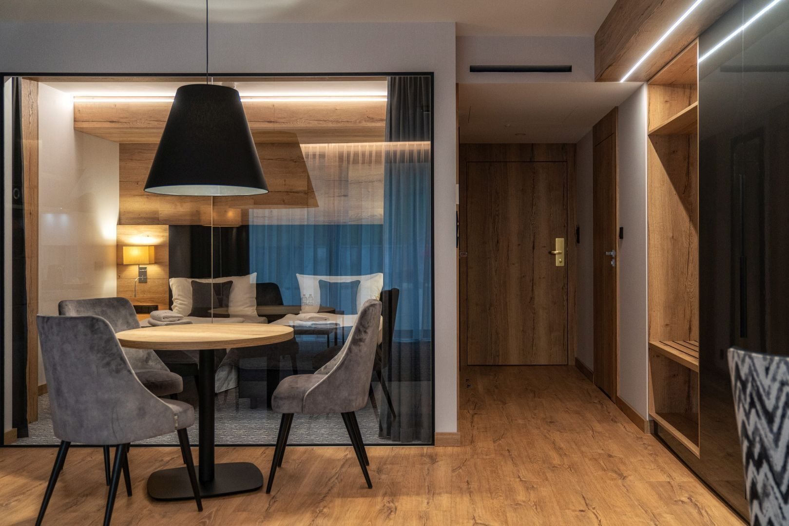 Une chambre d’hôtel moderne avec une table à manger ronde, des chaises grises à revêtement en tissu, une suspension noire et un parquet en bois.