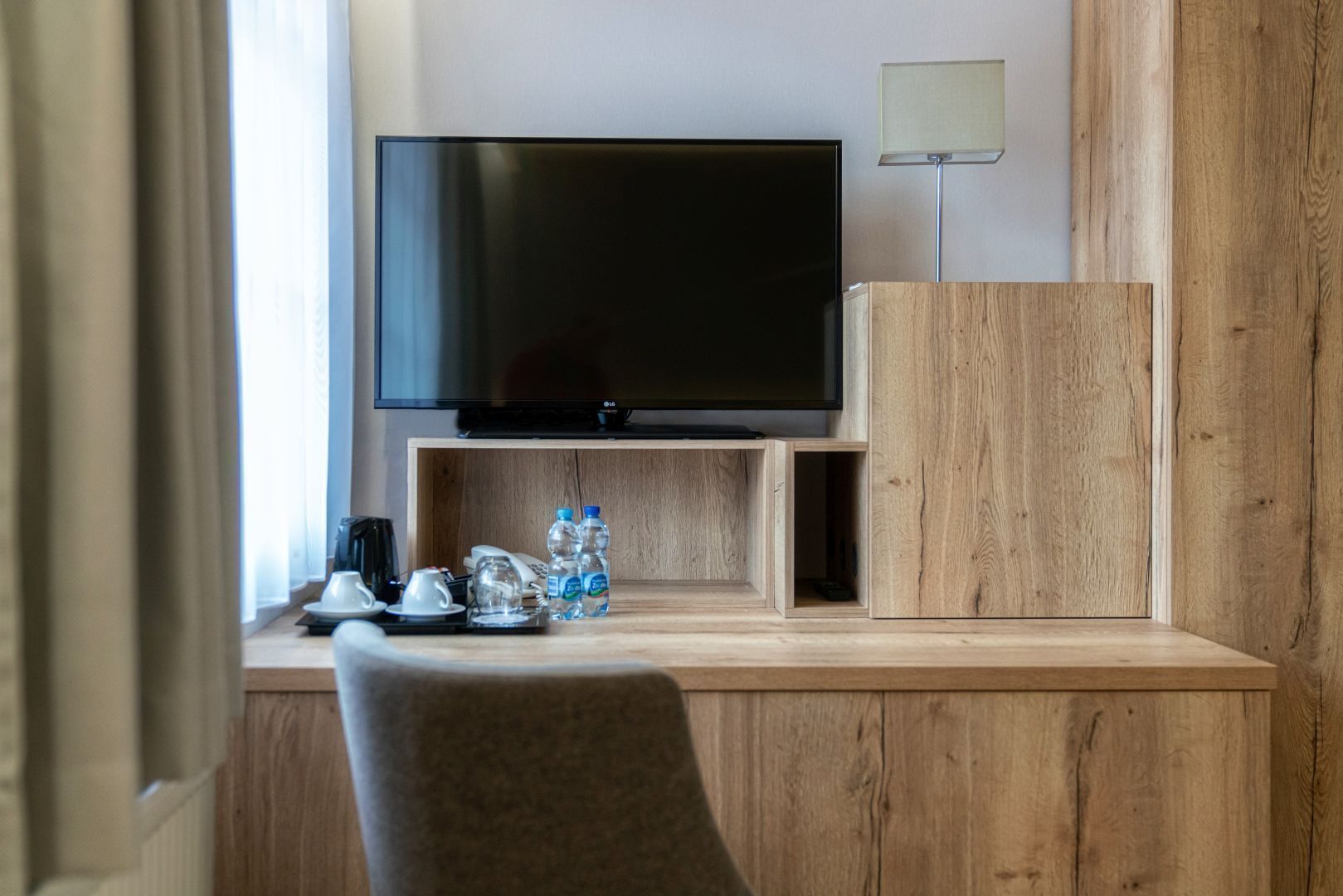 Habitación de hotel con una televisión de pantalla plana en una pared de madera, un mueble bajo y una mesita de noche con una lámpara y dos botellas de agua.