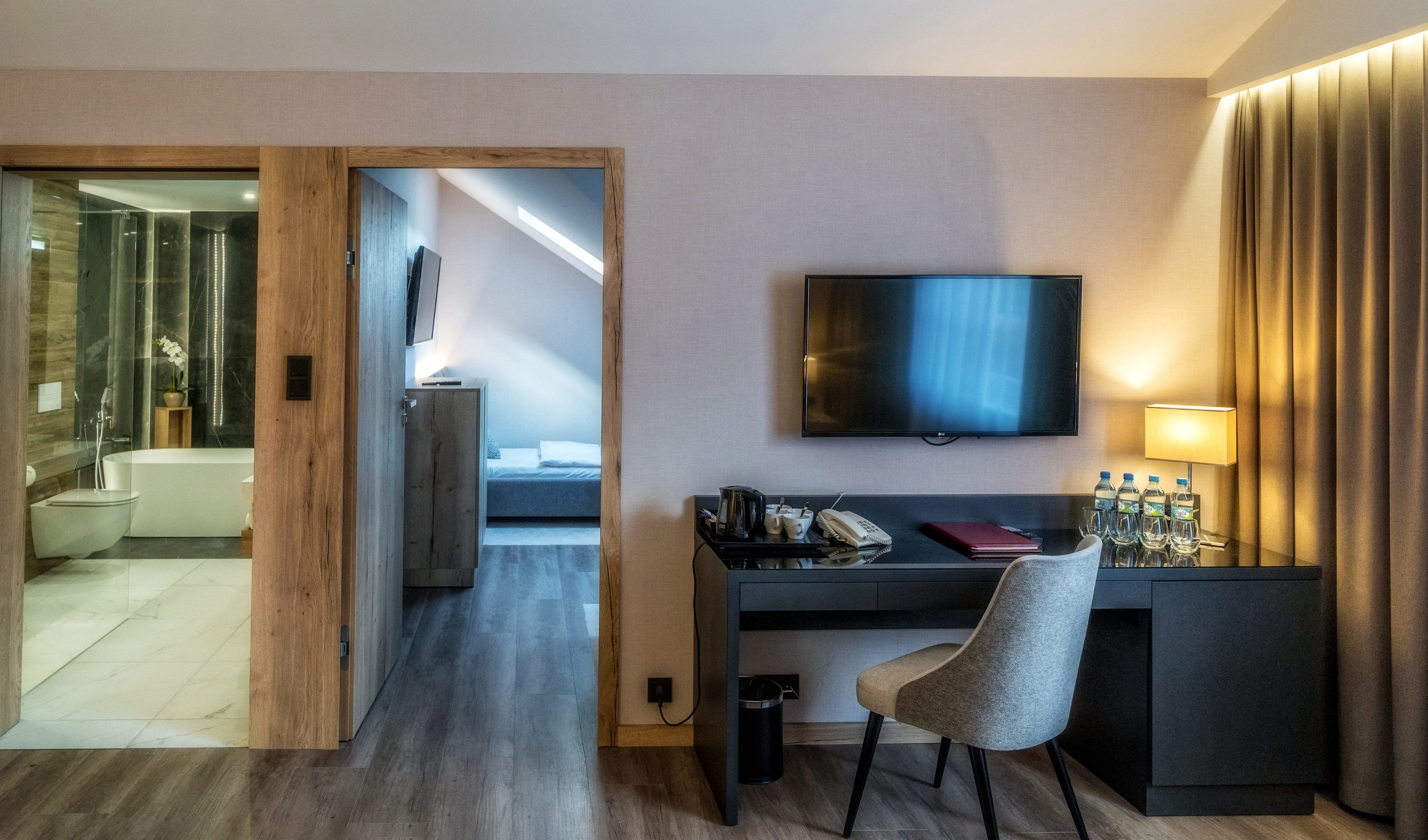 A hotel room with a wooden door leading to a bathroom, a desk with a chair, a flat-screen TV, and a bedside table with a lamp and water bottles.