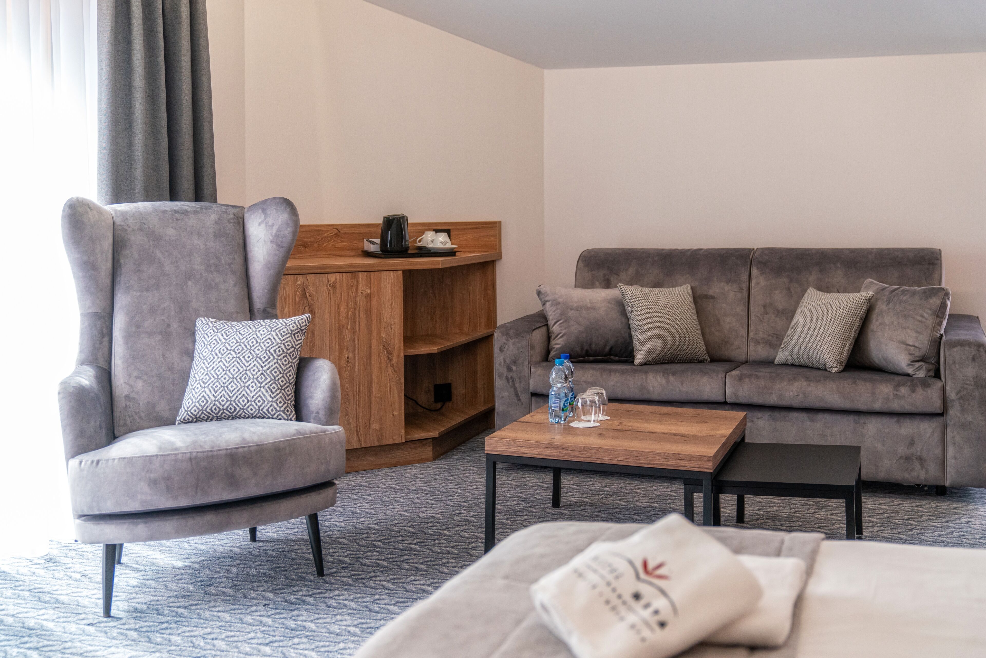 A modern hotel room with a grey armchair, a grey sofa, a wooden coffee table, and a small wooden side table with a kettle and cups.