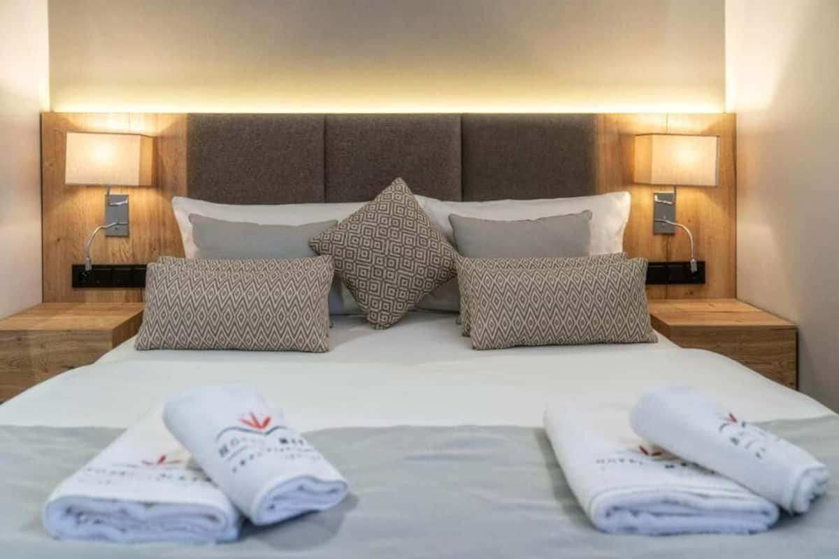 Una cama bien tendida con almohadillas estampadas, dos lámparas de noche y toallas enrolladas sobre la cama.