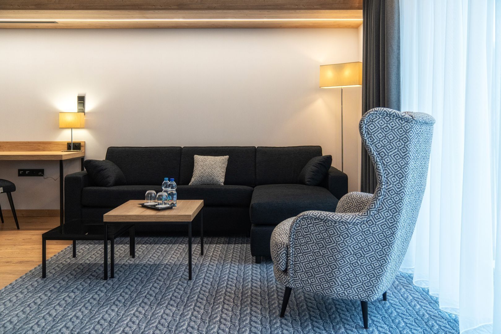 Ett modernt vardagsrum med en soffa, en mönstrad fåtölj, ett soffbord med vattenflaskor och en lampa.