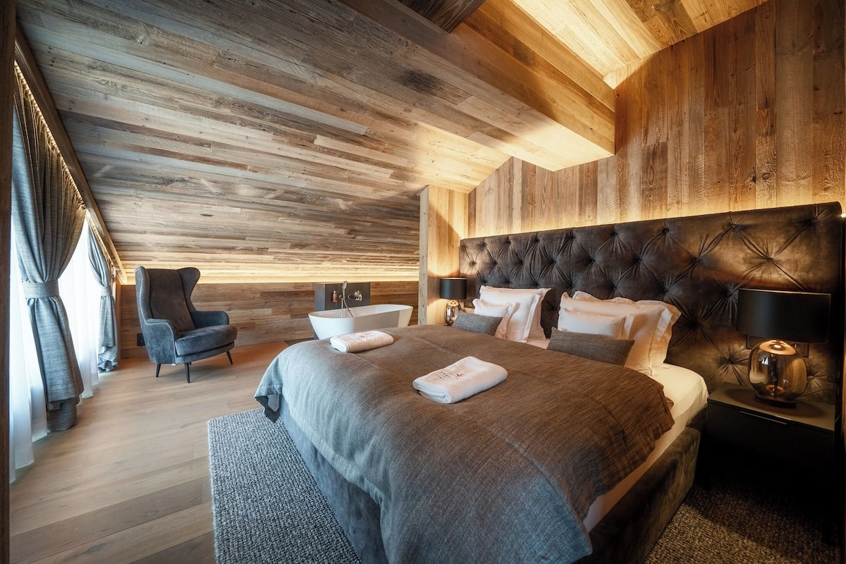 Un dormitorio moderno con techo de madera, una cama grande con cabecera capitonada, una mesita de noche con lámpara y una silla.