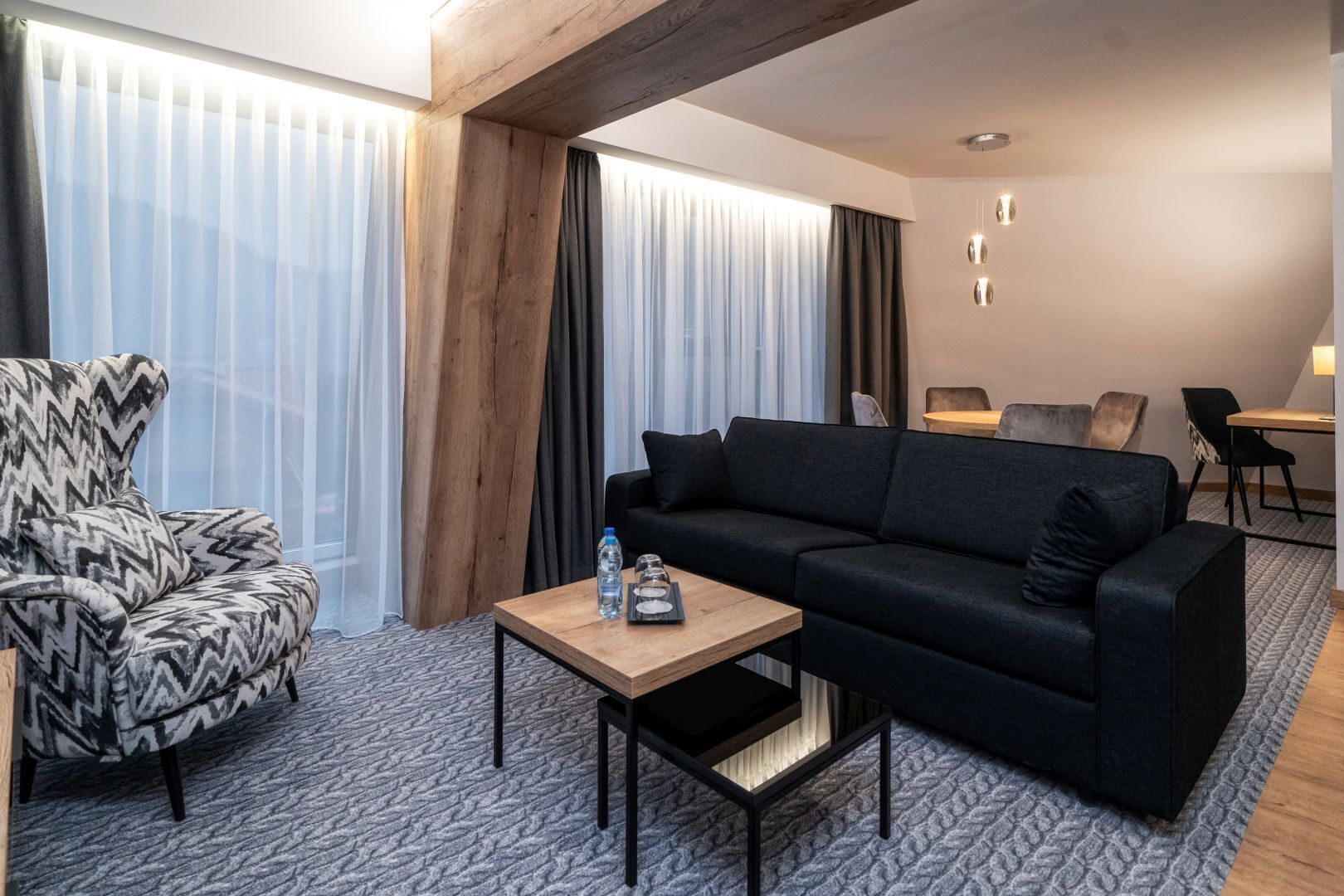 A modern hotel room with a black sofa, patterned armchair, and a small wooden table with a water bottle and glass.