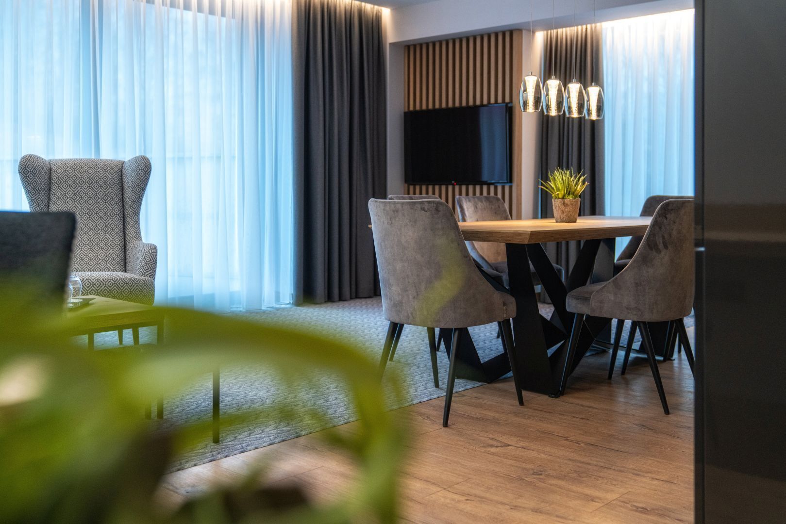 Una habitación de hotel moderna con zona de comedor, televisor grande y planta en maceta.