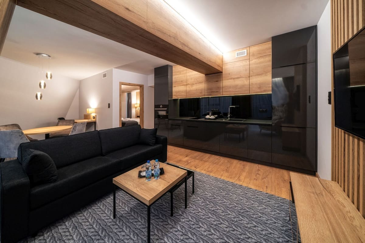 Una sala de estar moderna con un sofá negro, una mesa de centro de madera y una cocina con armarios oscuros y un refrigerador negro.
