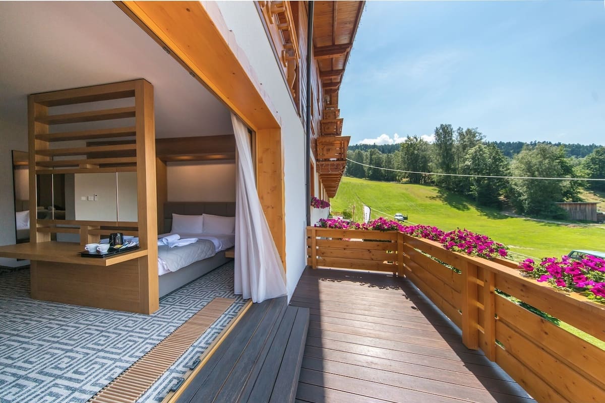 Un balcón con piso de madera, una cama con ropa blanca y vistas a un paisaje verde.