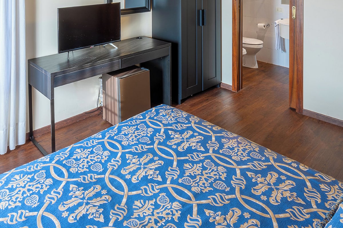 A room with a bed, a desk, a television, a wardrobe, and a bathroom visible through an open door.