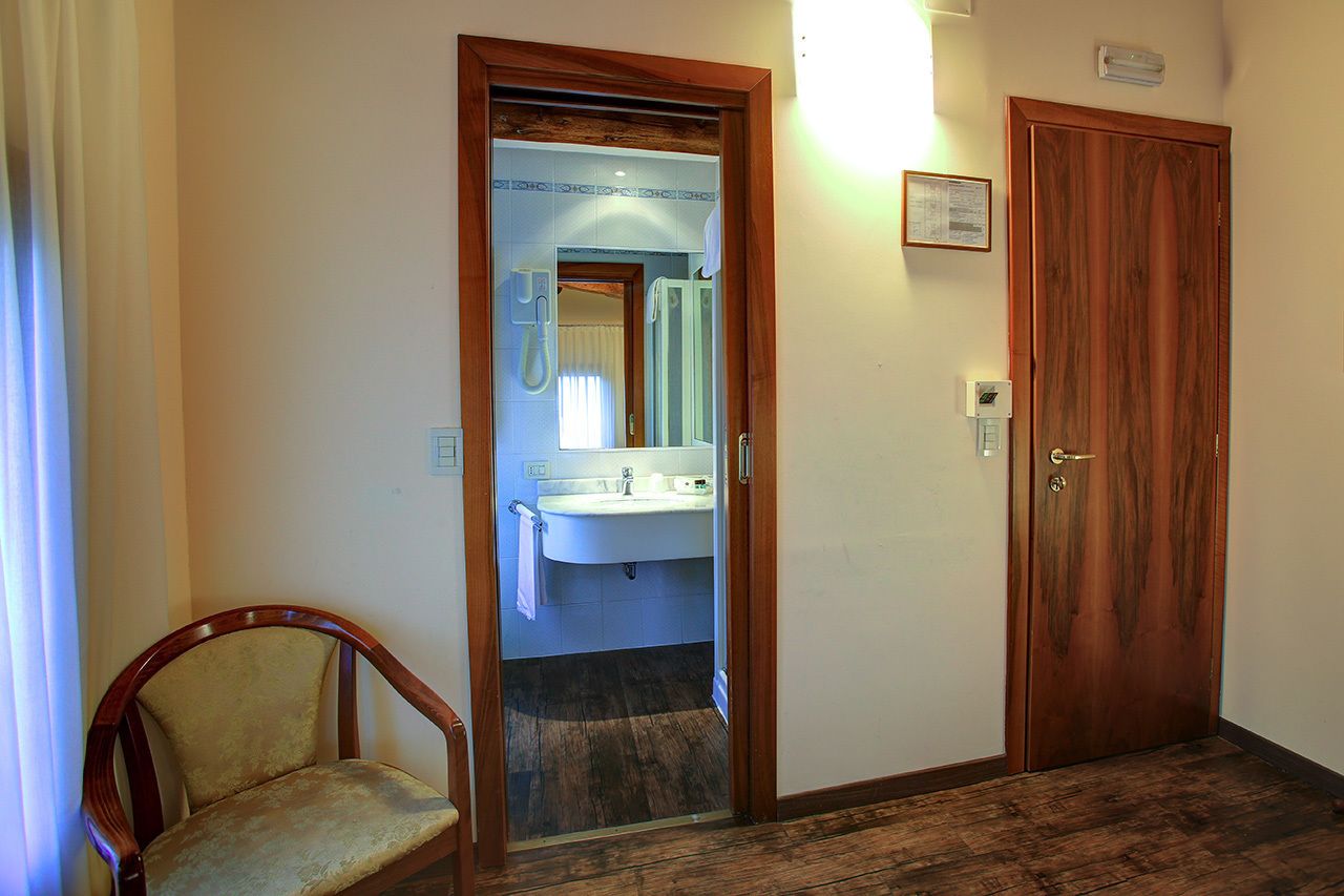 Quarto de hotel com porta de madeira, banheiro pequeno com pia e espelho, e uma cadeira de madeira.