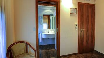 Quarto de hotel com porta de madeira, banheiro pequeno com pia e espelho, e uma cadeira de madeira.